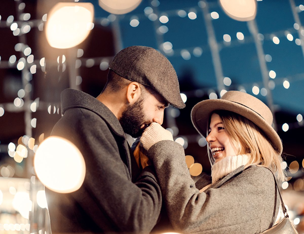 Romantic Christmas lovers have romance on Christmas eve. A young gentleman standing outdoors with his girlfriend and kissing her hand on New Year's eve. The couple is surrounded by Christmas lights.